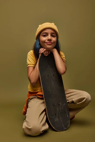 Pleased and stylish preadolescent child with dyed hair wearing urban outfit and yellow hat while looking at camera and holding skateboard on khaki background, girl with cool street style look — Stock Photo