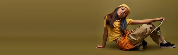 Criança pré-adolescente elegante com cabelo tingido usando chapéu amarelo e roupas urbanas posando e olhando para a câmera enquanto sentado no skate no fundo cáqui, menina no conceito de streetwear urbano, banner — Fotografia de Stock