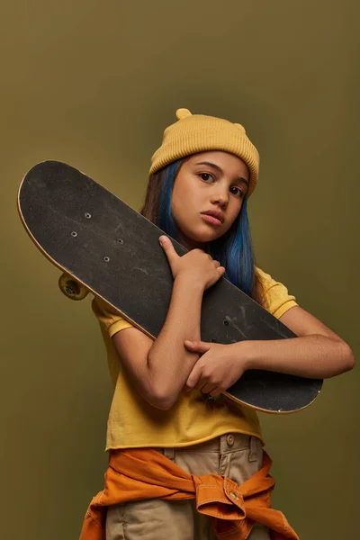 Porträt eines stilvollen vorpubertären Mädchens mit gelbem Hut und urbanem Outfit, das in die Kamera blickt, während es Skateboard hält und isoliert auf Khaki posiert, Mädchen in urbanem Streetwear-Konzept — Stockfoto