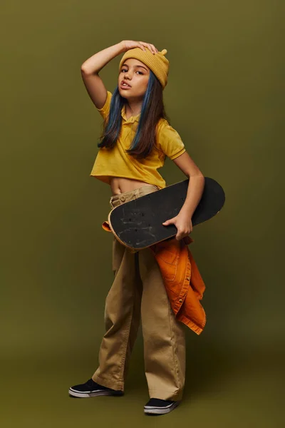 Full length of trendy preteen girl with dyed hair wearing urban outfit and holding skateboard while grimacing and looking at camera on khaki background, girl in urban streetwear concept — Stock Photo