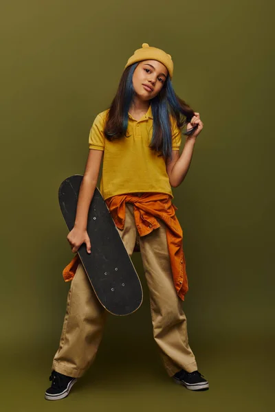 Selbstbewusstes und modisches Mädchen mit gefärbten Haaren, das Hut und urbanes Outfit trägt und Skateboard auf khakifarbenem Hintergrund hält, Mädchen in urbanem Streetwear-Konzept — Stockfoto