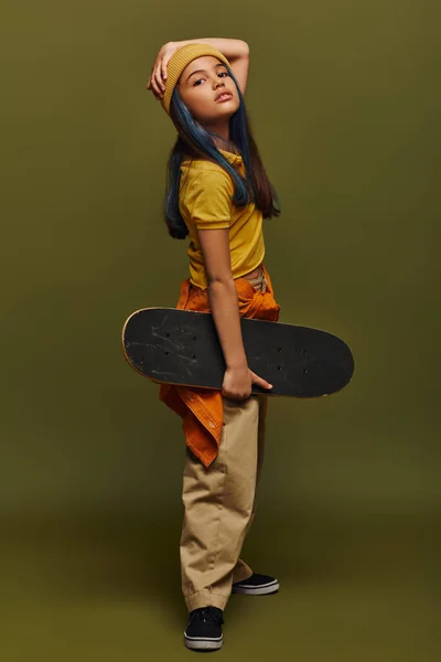 Volle Länge des trendigen und selbstbewussten vorpubertären Kindes mit farbigen Haaren, das Hut und urbanes Outfit trägt und Skateboard hält, während es auf khakifarbenem Hintergrund steht, Mädchen im urbanen Streetwear-Konzept — Stockfoto