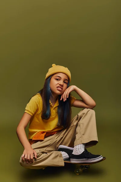 Disgusted and fashionable preteen girl with dyed hair wearing yellow hat and urban outfit while looking at camera and sitting on skateboard on khaki background, girl in urban streetwear concept — Stock Photo