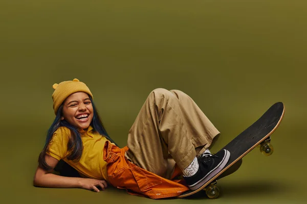 Excited and cheerful preteen girl with dyed hair wearing trendy clothes and hat while lying near skateboard and looking at camera on khaki background, girl in urban streetwear concept — Stock Photo