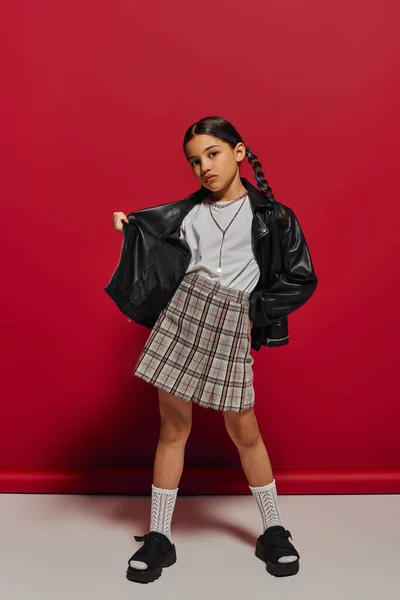 Full length of fashionable preadolescent girl with hairstyle posing in leather jacket and checkered skirt and looking at camera while standing on red background, stylish preteen outfit concept — Stock Photo