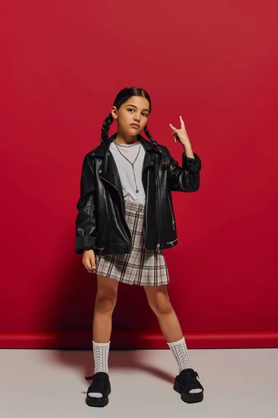 Full length of fashionable preteen girl with hairstyle wearing leather jacket and plaid skirt while showing rock sign at camera and standing on red background, stylish preteen outfit concept — Stock Photo
