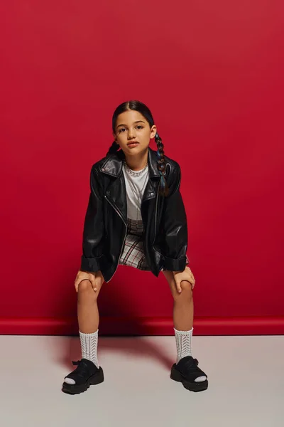 Full length of fashionable and confident preteen girl in leather jacket and plaid skirt looking at camera while posing and standing on red background, stylish preteen outfit concept — Stock Photo