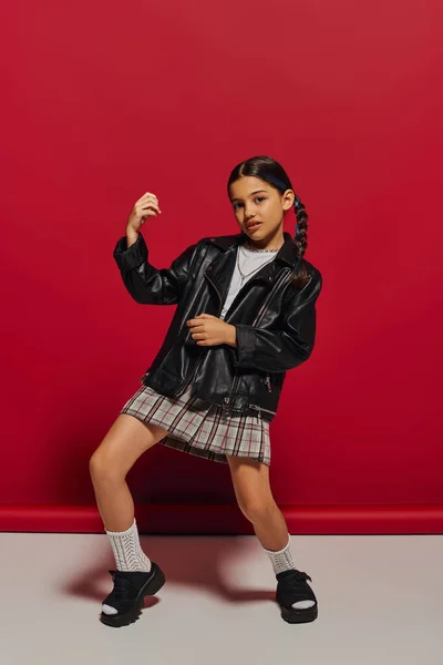 Volle Länge des trendigen und selbstbewussten vorpubertären Mädchens mit Frisur posiert in Lederjacke und kariertem Rock auf rotem Hintergrund, stilvolles Preteen-Outfit-Konzept — Stockfoto