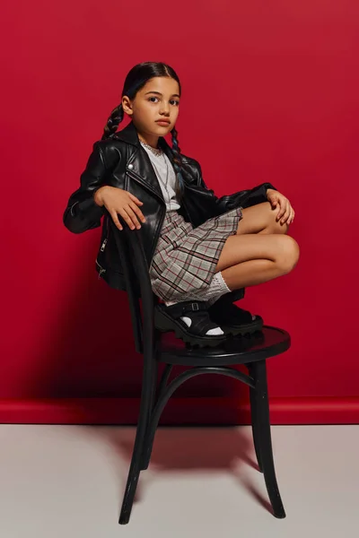 Fashionable preadolescent girl with hairstyle wearing leather jacket and plaid skirt while looking at camera and posing on chair on red background, stylish preteen outfit concept — Stock Photo