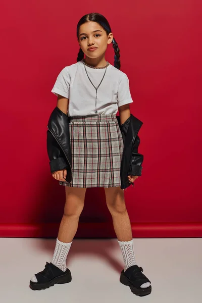 Full length of trendy brunette preteen girl with hairstyle posing in plaid skirt and leather jacket while looking at camera and standing on red background, stylish preteen outfit concept — Stock Photo