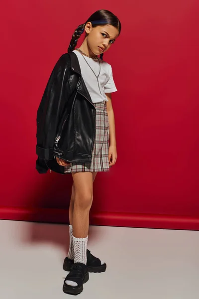 Серьёзная и предподростковая стильная девушка с прической, смотрящая в камеру, позируя в кожаной куртке и клетчатой юбке и стоя на красном фоне, стильная детская одежда — стоковое фото