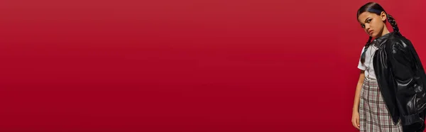 Menina da moda pré-adolescente descontente com penteado vestindo jaqueta de couro e saia xadrez enquanto olha para a câmera e de pé isolado no vermelho, conceito de roupa pré-adolescente elegante, banner — Fotografia de Stock