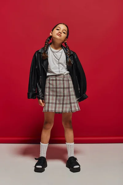 Full length of confident and fashionable preteen girl with hairstyle wearing leather jacket and plaid skirt while standing and posing on red background, stylish preteen outfit concept — Stock Photo