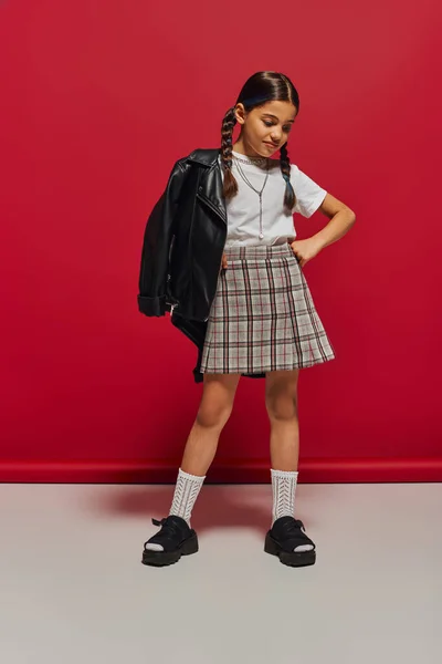 Trendiges brünettes Preteen-Mädchen mit Frisur posiert in Lederjacke und kariertem Rock, hält die Hand an der Hüfte und steht auf rotem Hintergrund, stilvolles Preteen-Outfit-Konzept — Stockfoto