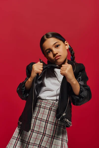 Porträt eines modischen vorpubertären Mädchens mit Frisur, das in kariertem Rock posiert, während es eine Lederjacke in der Hand hält und isoliert auf rotes, stilvolles Preteen-Outfit blickt — Stockfoto