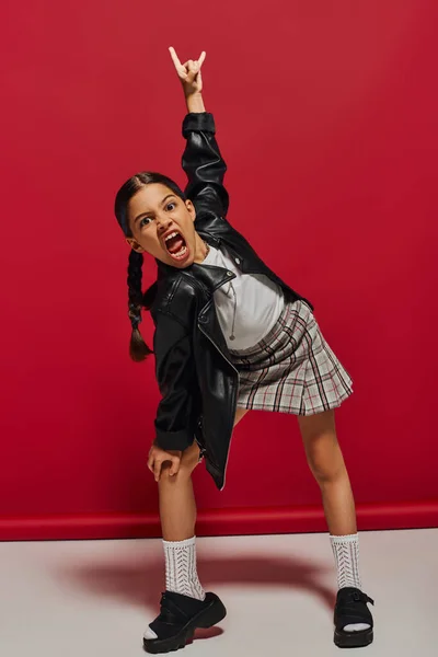 Mad and trendy preteen girl with hairstyle wearing leather jacket and plaid skirt looking at camera and showing rock gesture while standing on red background, stylish preteen outfit concept — Stock Photo