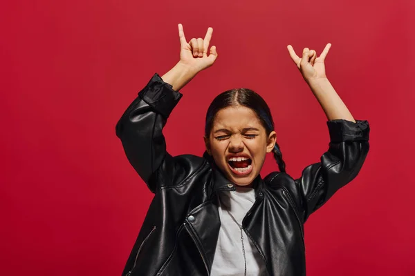 Excited and fashionable preteen girl with modern hairstyle closing eyes while posing in leather jacket and showing rock gesture isolated on red, girl with cool and contemporary look — Stock Photo