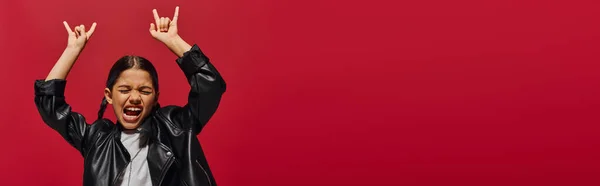 Menina pré-adolescente animado e elegante com penteado moderno usando jaqueta de couro e mostrando gesto rock isolado no vermelho, menina com olhar fresco e contemporâneo, banner com espaço de cópia — Fotografia de Stock