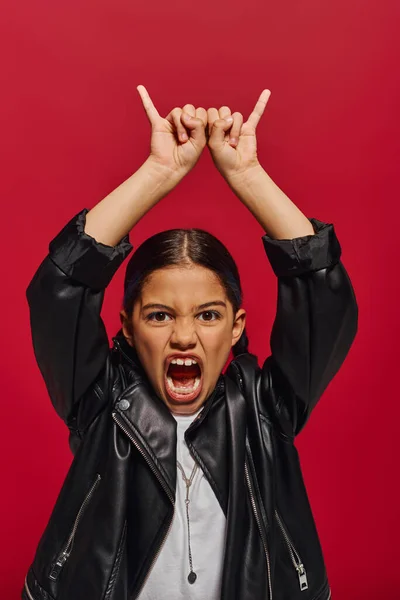 Portrait of mad and fashionable preadolescent girl in leather jacket screaming at camera and showing rock gesture while standing isolated on red, girl with cool and contemporary look — Stock Photo