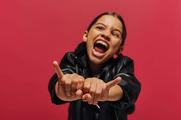 Menina pré-adolescente desfocada e louca em jaqueta de couro elegante mostrando gesto de rock na câmera enquanto em pé e posando isolado no vermelho, menina com olhar fresco e contemporâneo — Fotografia de Stock