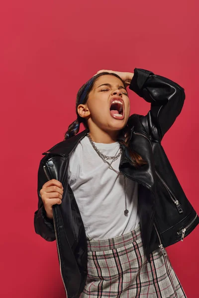 Angry and fashionable preteen girl with hairstyle screaming and touching head while posing in leather jacket and standing isolated on red, hairstyle and trendy accessories concept — Stock Photo