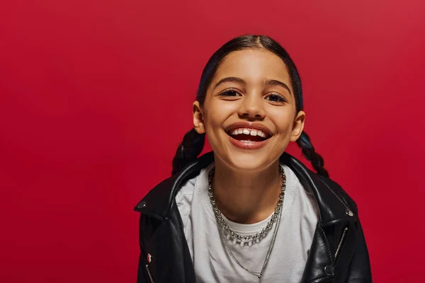 Retrato de menina pré-adolescente satisfeito com penteado vestindo jaqueta de couro elegante e olhando para a câmera enquanto posando isolado no vermelho, penteado e conceito de acessórios da moda — Fotografia de Stock