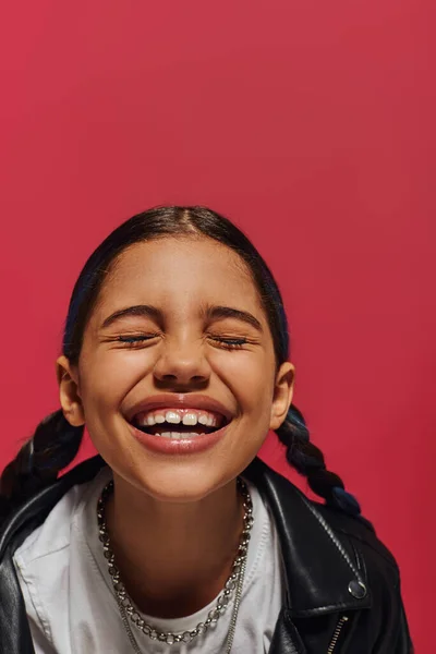 Porträt eines lächelnden Frühchens mit Frisur, die Halsketten und Lederjacke trägt, während sie mit geschlossenen Augen steht und isoliert auf Rot posiert, Frisur und trendiges Accessoire-Konzept — Stockfoto