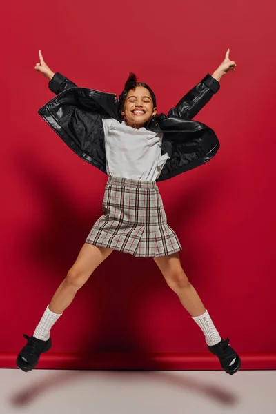 Full length of cheerful and fashionable preteen girl in plaid skirt and leather jacket while jumping and pointing with fingers on red background, hairstyle and trendy accessories concept — Stock Photo