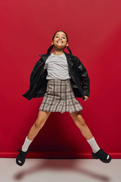 Full length of cheerful preteen kid in stylish leather jacket and plaid skirt looking at camera while jumping and having fun on red background, hairstyle and trendy accessories concept — Stock Photo