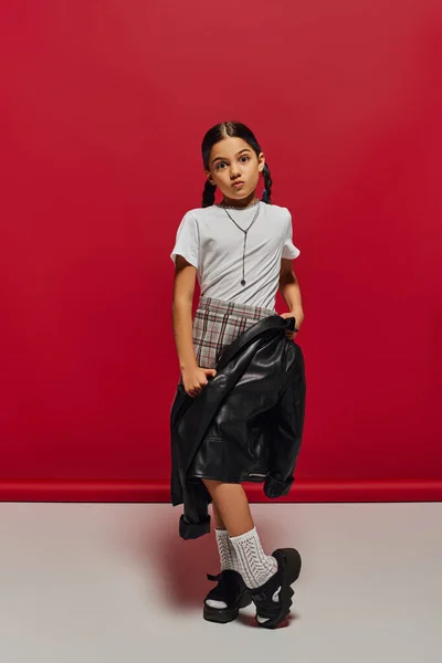 Preadolescent and fashionable girl with hairstyle wearing plaid skirt and holding leather jacket while looking at camera and posing on red background, hairstyle and trendy accessories concept — Stock Photo