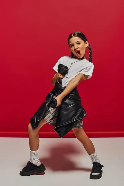 Excited and trendy preteen girl with hairstyle wearing t-shirt and plaid skirt while holding leather jacket and standing on red background, hairstyle and trendy accessories concept — Stock Photo
