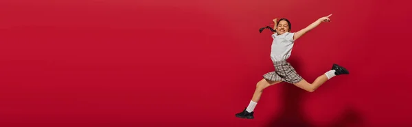 Позитивна і стильна дівчина-підліток у білій футболці і плетеній спідниці стрибає і вказує пальцем на червоному тлі, зачіска і концепція модних аксесуарів, банер — стокове фото