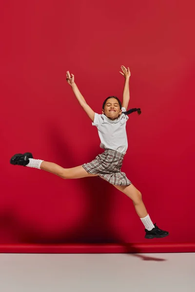 Garota pré-adolescente animado e alegre em t-shirt branca e elegante saia quadriculada pulando e se divertindo enquanto posando no fundo vermelho, penteado e conceito de acessórios na moda — Fotografia de Stock