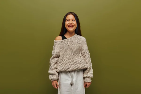 Gleeful and stylish brunette preteen girl with dyed hair wearing knitted sweater and looking away while standing and posing isolated on green, relaxed autumn vibes concept — Stock Photo