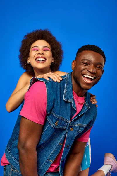 Cheerful young african american woman with bold makeup hugging best friend in stylish outfit and having fun while standing together isolated on blue, best friends in matching outfits, friendship — Stock Photo