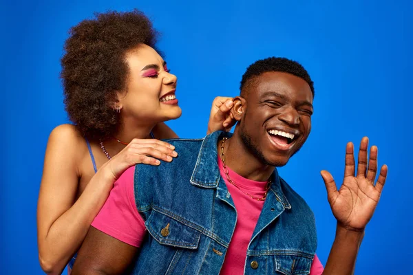 Позитивна молода афроамериканська жінка з сміливим макіяжем, що торкається вуха стильного найкращого друга в літньому вбранні, розважаючись і стоячи ізольовано на синьому, найкращі друзі в відповідному вбранні — стокове фото
