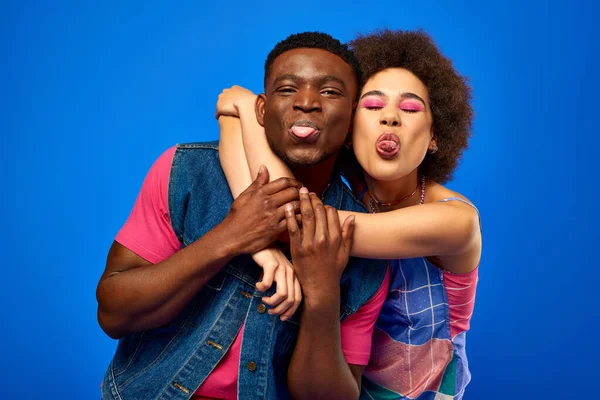 Positive junge afrikanisch-amerikanische beste Freunde in stylischen hellen Sommeroutfits, die die Zunge herausstrecken und Spaß haben, während sie isoliert auf blauem Grund stehen, beste Freunde in passenden Outfits — Stockfoto
