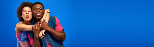 Giovani afroamericani migliori amici in abiti estivi luminosi e alla moda che abbracciano e sporgono lingue mentre stanno insieme isolati su blu, banner, migliori amici in abiti coordinati — Foto stock