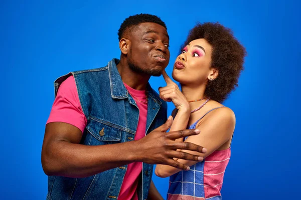 Young african american woman with bold makeup touching cheek of best friend in stylish summer outfit while standing together and having fun isolated on blue, best friends in matching outfits — Stock Photo