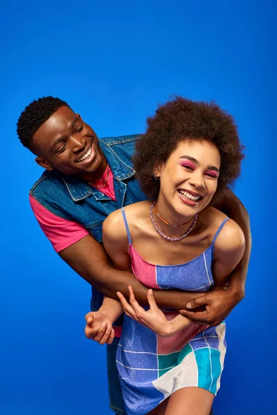 Homem americano africano positivo em roupa de verão brilhante abraçando o melhor amigo elegante com maquiagem ousada e sundress enquanto estão juntos isolados em azul, melhores amigos em roupas correspondentes — Fotografia de Stock