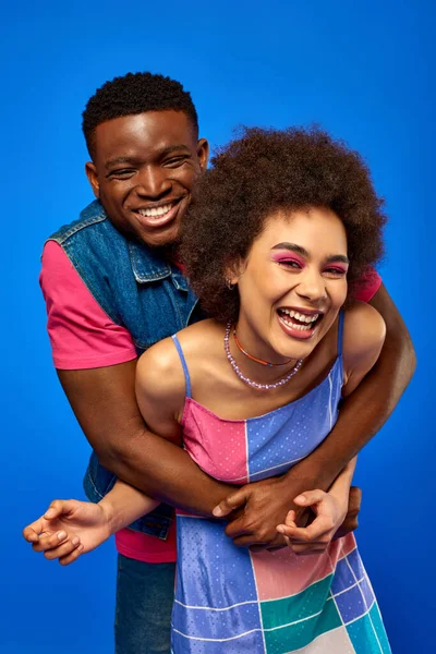 Cheerful young african american man in bright summer outfit hugging stylish best friend with bold makeup and sundress while standing isolated on blue, best friends in matching outfits — Stock Photo