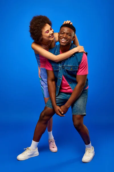 Comprimento total de positiva jovem afro-americana em sundress abraçando melhor amigo no verão roupa elegante, enquanto de pé e se divertindo no fundo azul, os melhores amigos se divertindo — Fotografia de Stock
