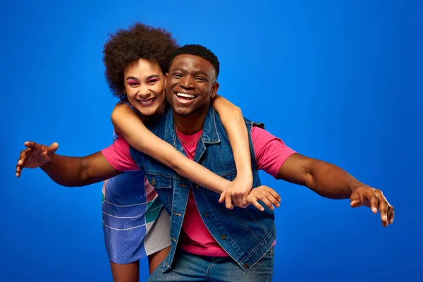 Весела молода афроамериканська жінка зі сміливим макіяжем і сарафанчиком обіймає модного найкращого друга в літньому вбранні, стоячи разом ізольовано на синьому, найкращі друзі добре проводять час — стокове фото