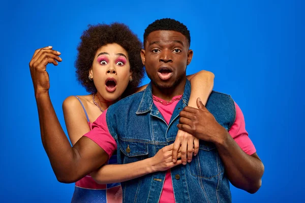 Jovem afro-americana chocada com maquiagem ousada abraçando melhor amiga em roupa de verão brilhante enquanto olha para a câmera e está isolada em azul, amigos da moda em roupas da moda — Fotografia de Stock