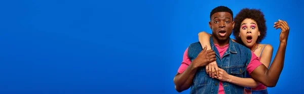 Scioccato giovane donna afroamericana con trucco audace abbracciando elegante migliore amico in abiti estivi e guardando la fotocamera isolata su blu, amici alla moda in abiti alla moda, banner — Foto stock