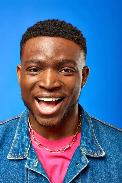 Portrait of cheerful young african american man with modern hairstyle wearing denim vest and looking at camera while standing and posing isolated on blue, trendy man showing summer style — Stock Photo