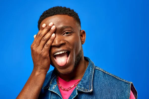 Excited young african american man in denim vest and bright t-shirt sticking out tongue and covering face while standing isolated on blue, trendy man showing summer style — Stock Photo