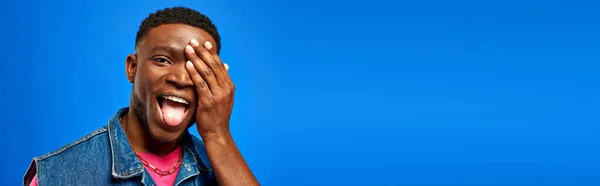 Emocionado jovem afro-americano com penteado moderno cobrindo rosto e salientando a língua enquanto posando em roupas de verão isolado em azul, homem da moda mostrando estilo de verão, banner — Fotografia de Stock