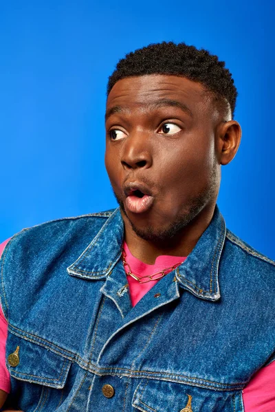 Portrait of shocked young african american man with modern hairstyle wearing summer outfit and looking away while standing and posing isolated on blue, trendy man showing summer style — Stock Photo