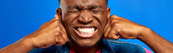 Agressivo jovem afro-americano com olhos fechados em roupas de verão de mãos dadas em punhos enquanto em pé e posando isolado em azul, homem da moda mostrando estilo de verão, banner — Fotografia de Stock